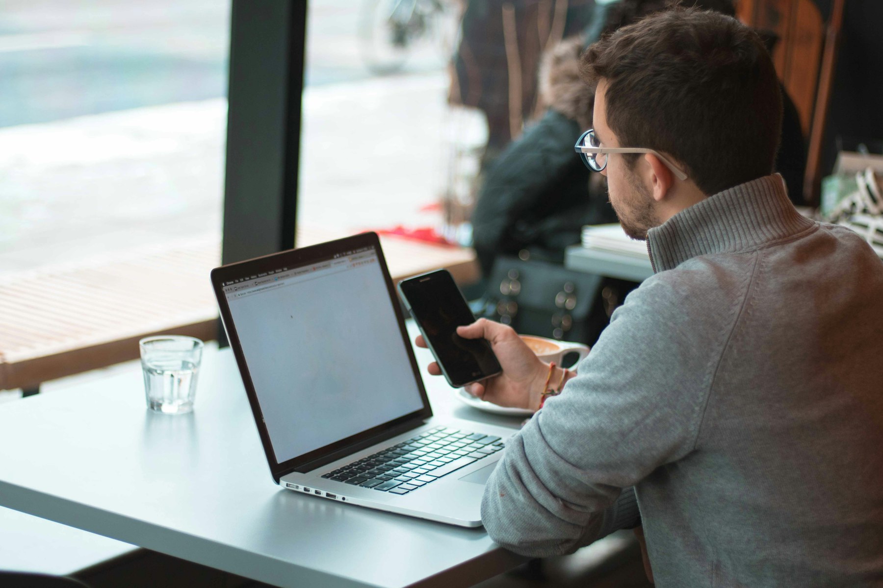 Man working remotely at a café table, checking his phone next to his laptop